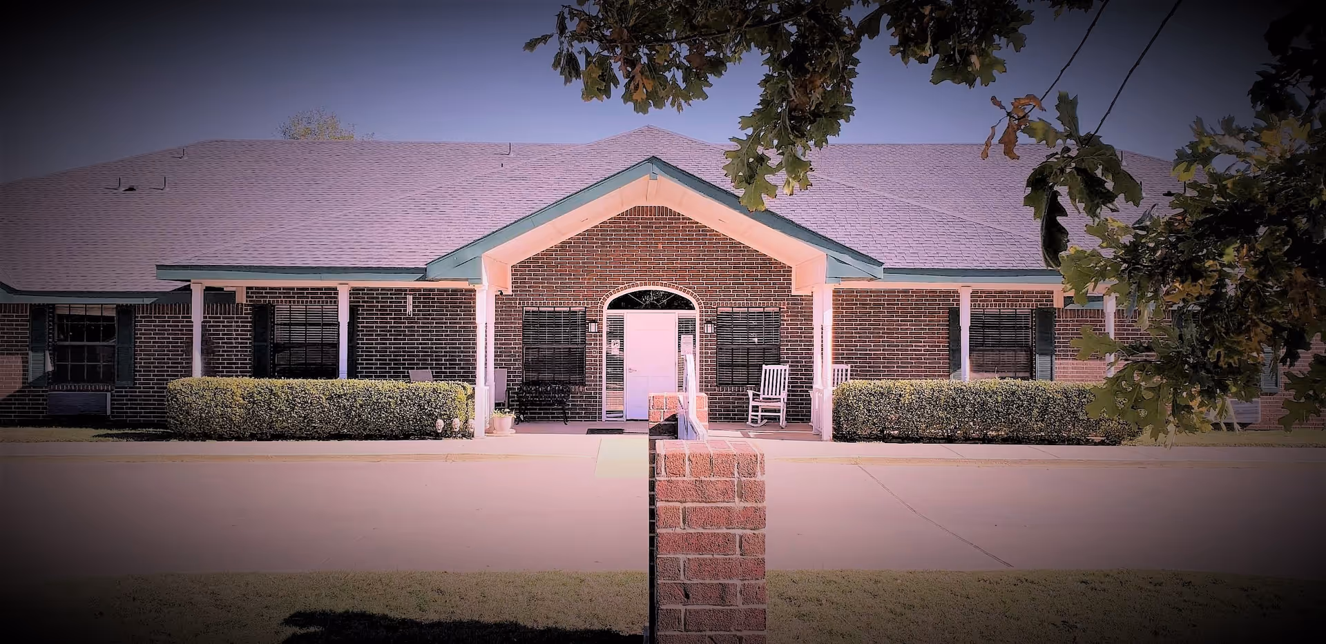 Front exterior view of a single-story brick building with a covered entrance supported by white columns. There are bushes along the front and two white rocking chairs on the porch. Tree branches frame the top of the image.