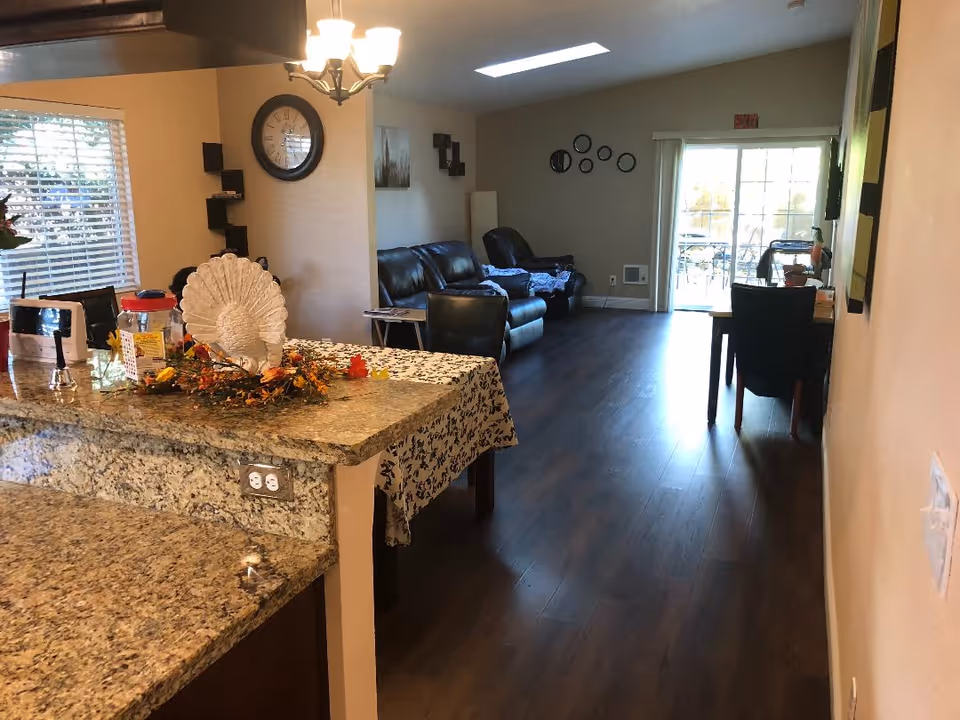Open-plan interior showing a granite kitchen island in the foreground, a dining table, and a living room with sofas leading to sliding glass doors.