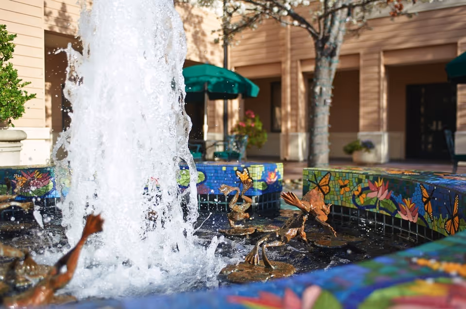 A colorful mosaic-tiled courtyard fountain with bronze frog sculptures and a central water jet in front of a building.