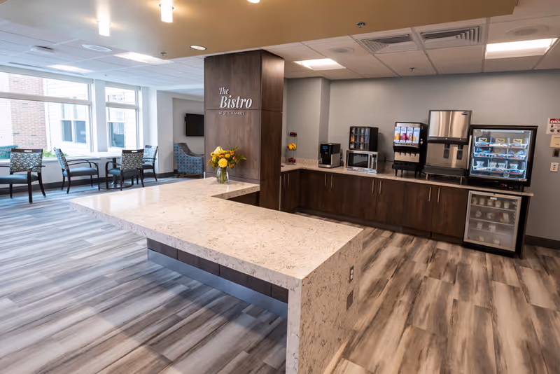 A modern dining area named The Bistro at Stella Maris featuring a large marble countertop island, dark wooden cabinets, a coffee machine, microwave, beverage dispensers, and a refrigerated display case. There are chairs and tables near large windows letting in natural light.