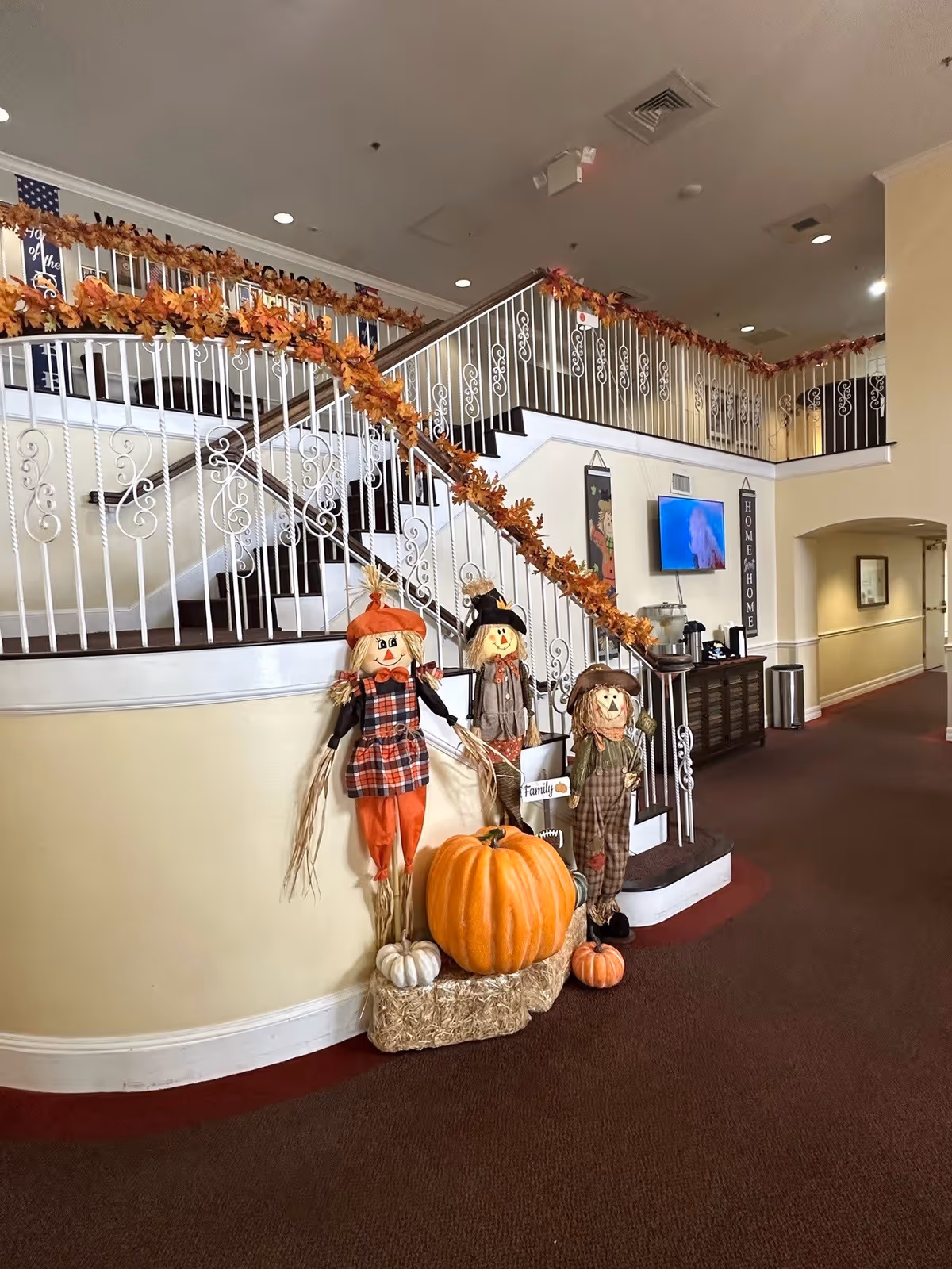 A decorated two-story lobby staircase with white ornate railings adorned with autumn garlands and fall decorations including scarecrows and pumpkins.