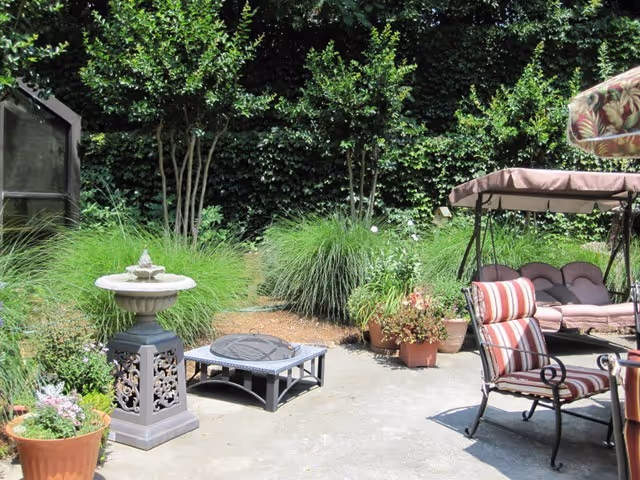 Outdoor patio area with a cushioned swing, a striped cushioned chair, a small table, potted plants, a decorative birdbath, and lush green bushes and trees in the background.