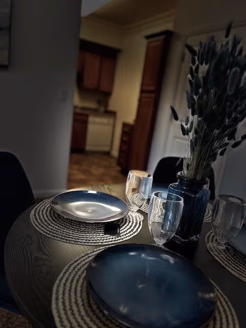 A close-up view of a dining table set with two dark plates, two clear drinking glasses, and a blue vase with dried flowers. The table has woven placemats and is situated near a kitchen area with wooden cabinets and a white dishwasher in the background.