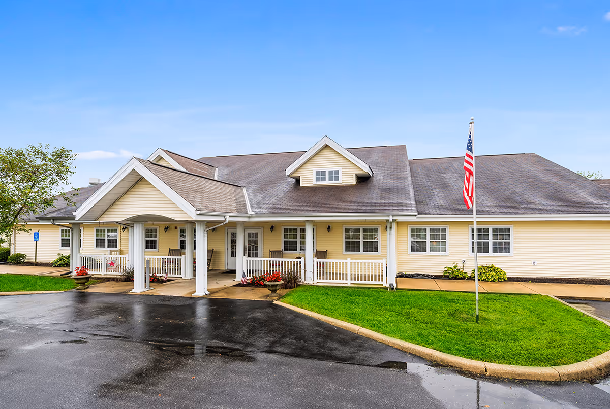 Front exterior of a single-story yellow senior living building with a covered entrance, white columns, an American flag, and a wet driveway.