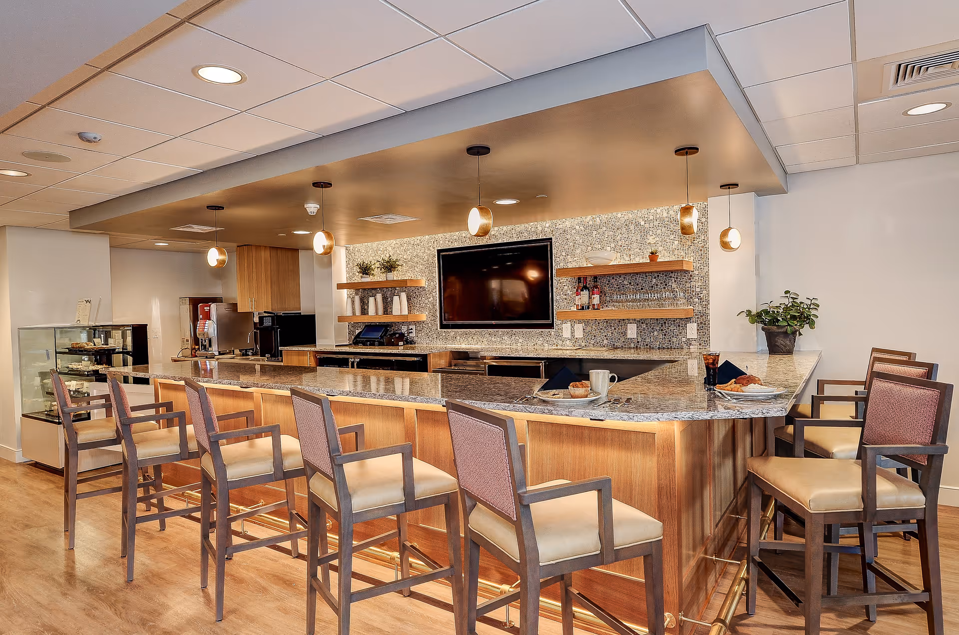 A modern kitchen and dining area with a large granite countertop island surrounded by wooden chairs with cushioned seats. The back wall features a mosaic tile backsplash, floating wooden shelves with decorative items, a mounted flat-screen TV, and pendant lights hanging from the ceiling. The floor is wooden, and there is a small display case on the left side.