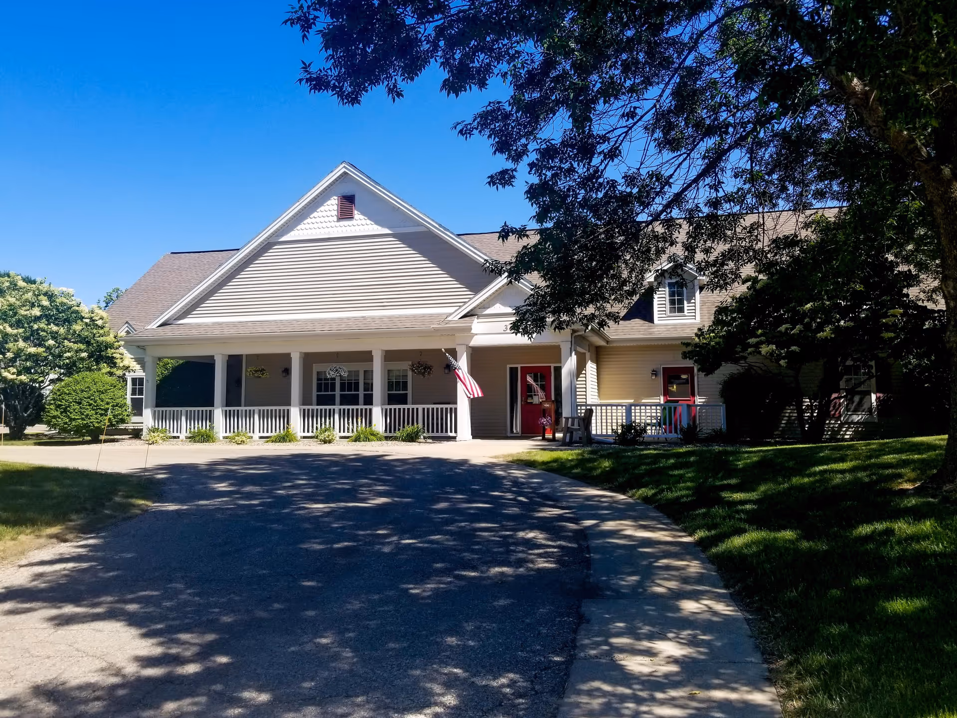 Front exterior of a single-story assisted living building with a covered porch, an American flag, and a curved driveway framed by trees.