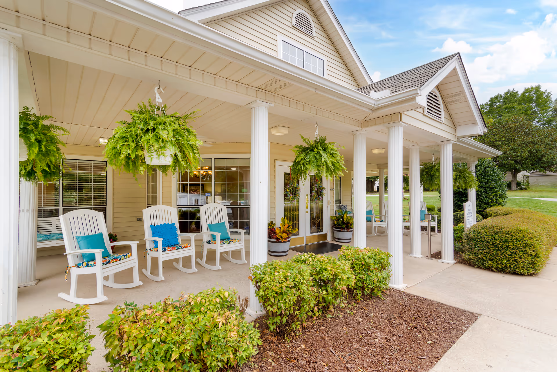 Covered porch area of a senior living facility with white rocking chairs adorned with colorful cushions, hanging green ferns, potted plants near the entrance, and white columns supporting the roof. The building exterior is beige with large windows and a double glass door entrance. There are shrubs and a walkway in front of the porch under a partly cloudy sky.