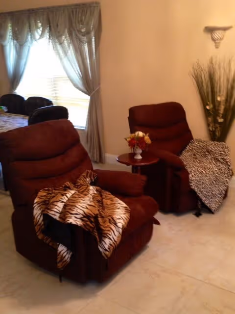 Two brown recliner chairs with animal print blankets draped over them in a living area. A small round table with a floral arrangement is placed between the chairs. In the background, there is a window with sheer curtains and a dining table with chairs. A tall decorative plant is positioned against the wall.