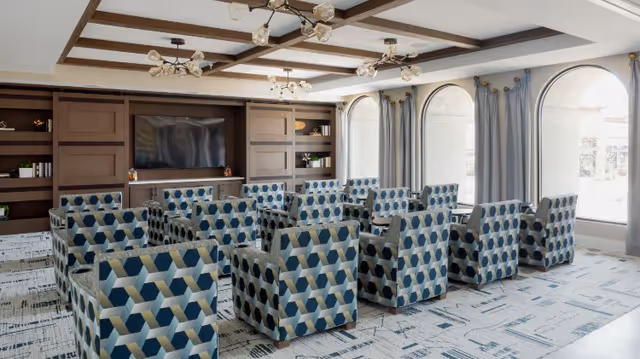 A bright common room with patterned armchairs arranged in rows facing a large TV and built-in shelving beneath a coffered ceiling and arched windows.