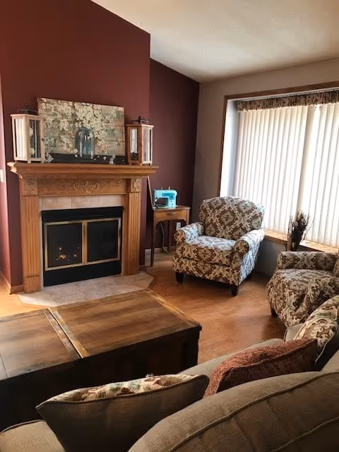 Cozy living room with a wooden fireplace on a burgundy accent wall, two patterned armchairs near a large window with vertical blinds, a wooden coffee table, and a small side table with a blue sewing machine on it.