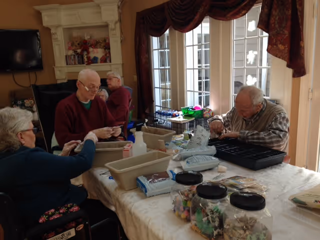 A group of elderly people sitting around a table engaged in a craft activity with various supplies like containers, bags, and jars filled with craft materials. The room has large windows with red curtains and a fireplace in the background.