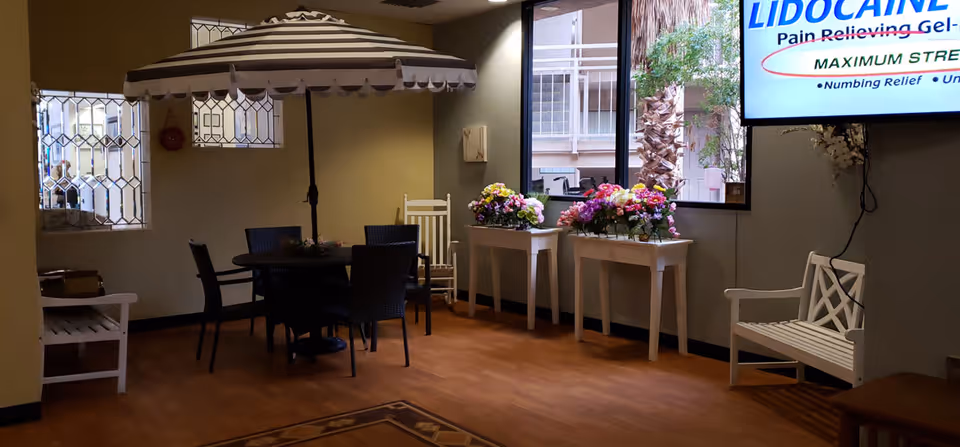 Indoor seating area with a round table and four chairs under a large striped umbrella. Two white benches are placed against the walls, and two white tables hold colorful flower arrangements near a window showing outdoor palm trees. A television screen on the wall displays an advertisement for Lidocaine pain-relieving gel.