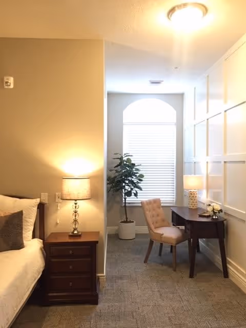 Well-lit bedroom corner with a bed and nightstand on the left and a small desk, chair, and potted plant by a window on the right.