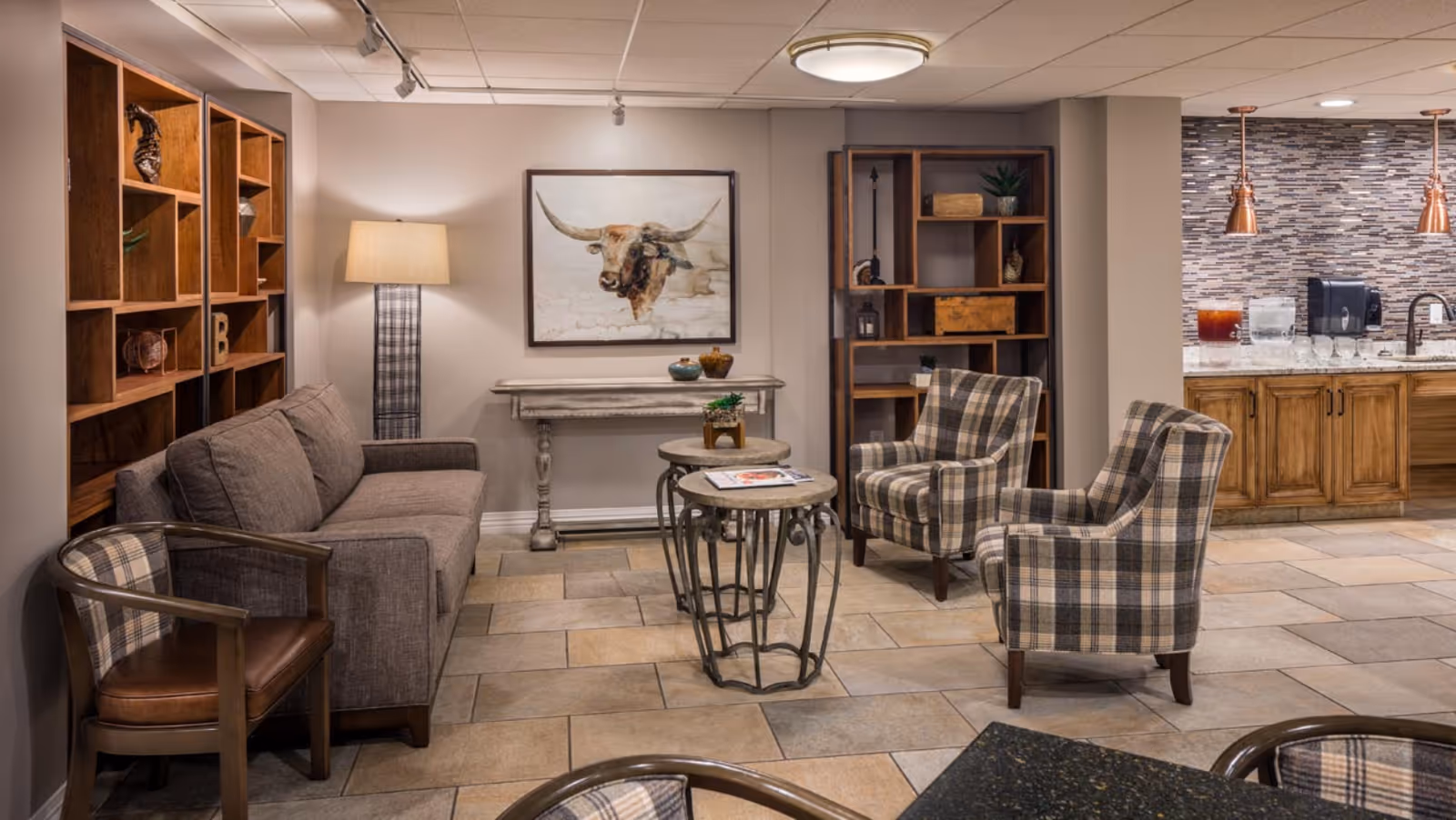 A cozy senior living common area featuring a gray sofa, two plaid armchairs, a wooden chair with a leather seat, and two round nesting tables. The room has a tiled floor, a wooden bookshelf with decorative items, a floor lamp, and a framed painting of a longhorn bull on the wall. In the background, there is a kitchenette area with wooden cabinets, a tiled backsplash, a sink, and beverage dispensers.