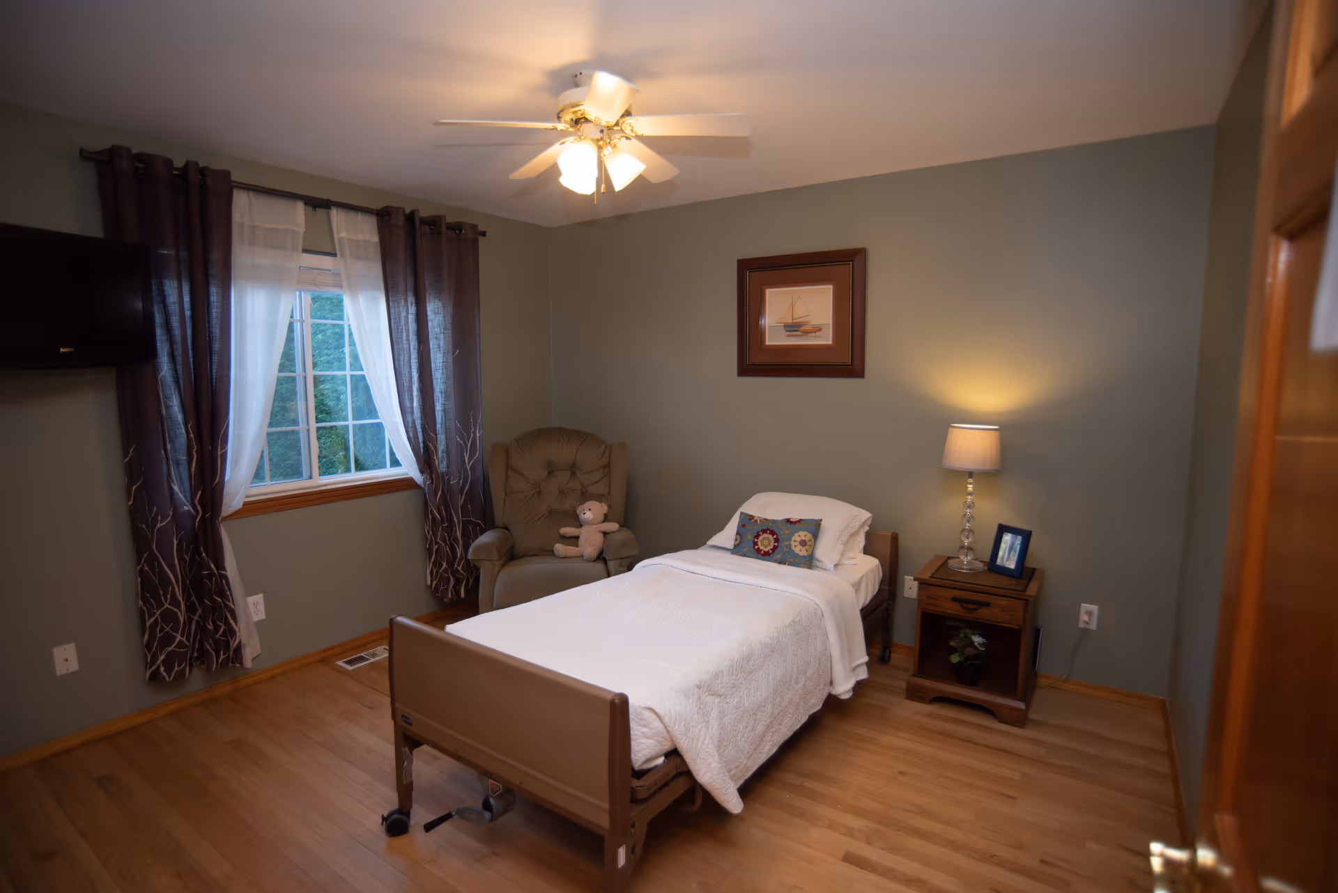 A cozy bedroom in a senior living facility with a single bed covered in white bedding, a decorative pillow, and a teddy bear on a cushioned armchair next to a window with dark and sheer curtains. A wooden nightstand with a lamp and a framed photo is beside the bed, and a ceiling fan with lights is overhead. The walls are painted green and there is a framed picture of a sailboat on the wall.