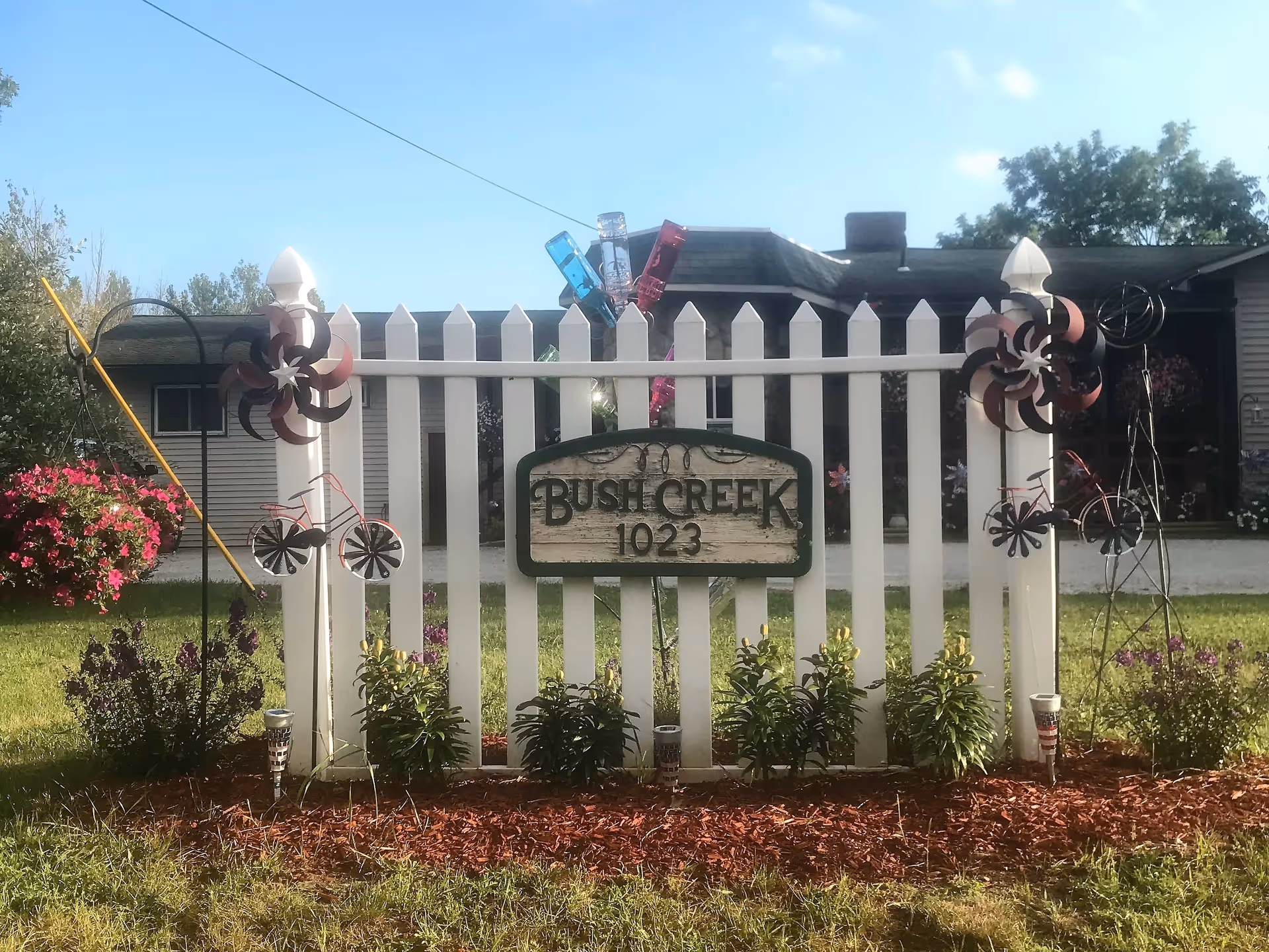 White picket fence with a wooden sign reading 'Bush Creek 1023' in front of a house. The fence is decorated with metal wind spinners shaped like bicycles and flowers, with plants and mulch at the base. The sky is clear and blue.