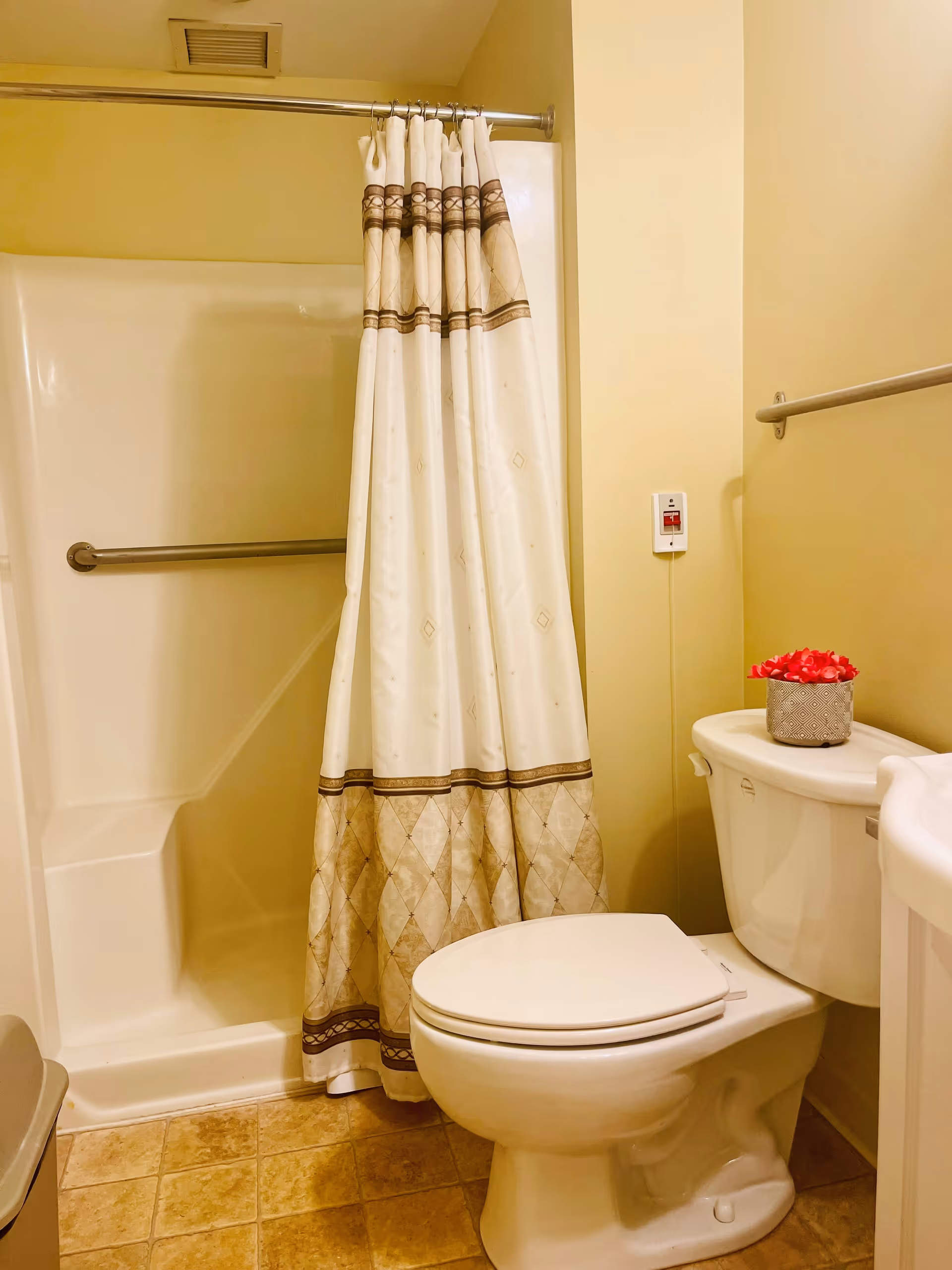 A small bathroom with a white toilet, a shower with a beige and brown patterned curtain, a grab bar inside the shower, a beige tiled floor, and a small decorative pot with red flowers on the toilet tank.