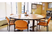 A round wooden table with six orange cushioned chairs arranged around it in a well-lit room with large windows. The table has some papers, a glass container, and a small decorative item on it. The room has light-colored walls and a bulletin board with notices on one side.