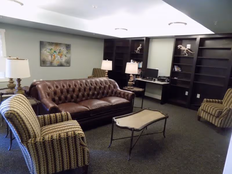 A cozy living room area featuring a brown leather tufted sofa, two striped armchairs, a glass-top coffee table, two table lamps on side tables, built-in dark wood bookshelves with dinosaur skeleton models, and a computer workstation.
