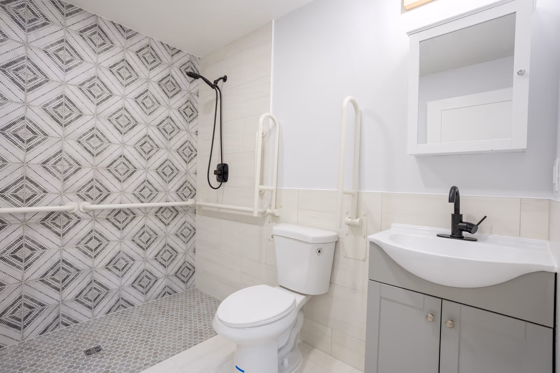 A modern bathroom with a walk-in shower featuring patterned tiles and a black showerhead. The bathroom includes a white toilet with grab bars on both sides and a white sink with a black faucet under a mirrored medicine cabinet.