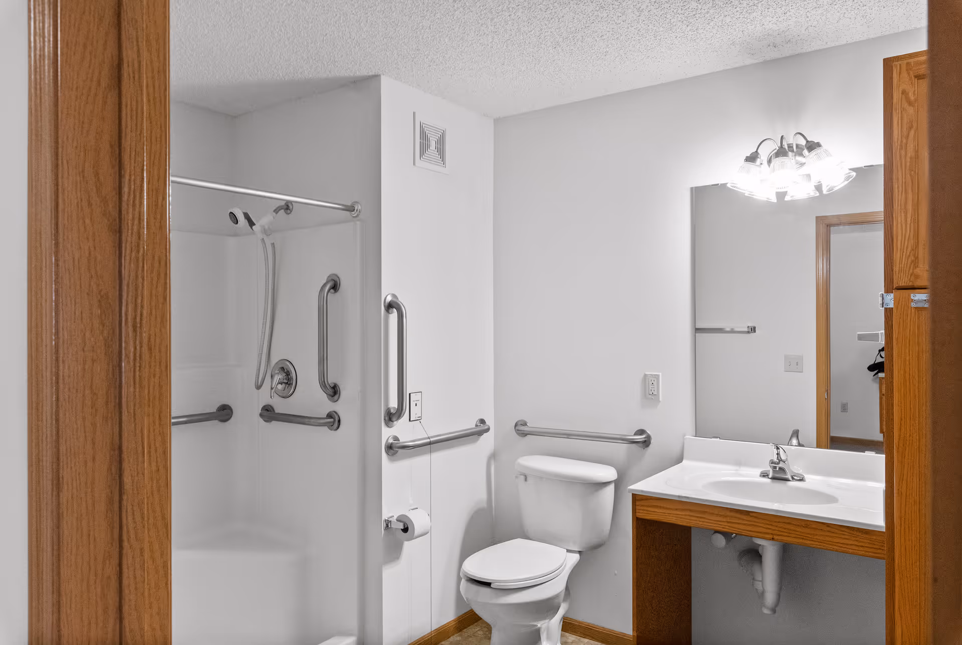 A clean and accessible bathroom with a white toilet, a white sink with a wooden vanity, a large mirror above the sink, and a shower with grab bars and a handheld showerhead. The walls are white, and the door frames are wooden.