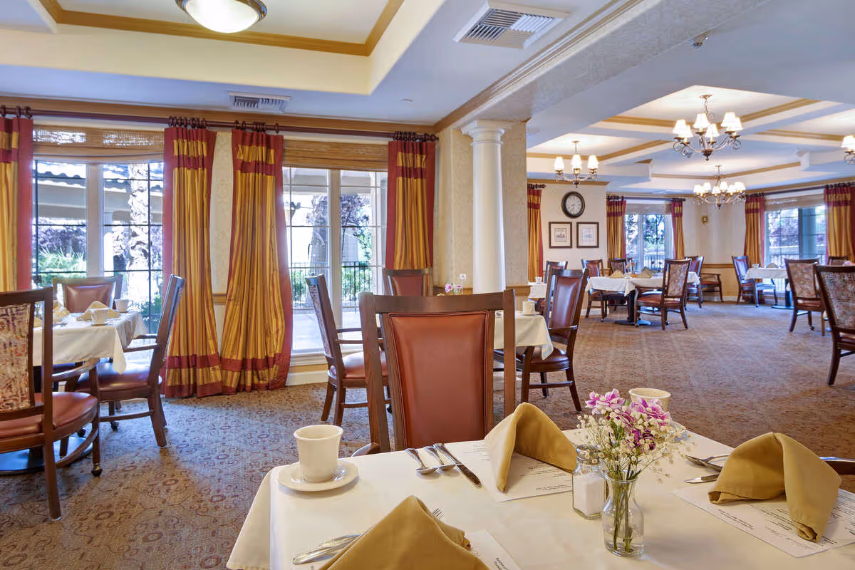 A spacious dining room with multiple tables set with white tablecloths, beige napkins, and cutlery. The room features large windows with red and gold curtains, carpeted floors, and elegant chandeliers hanging from the ceiling. There are chairs with wooden frames and leather backs around the tables, and a small vase with flowers is placed on the table in the foreground.