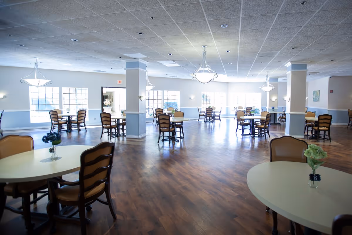 A spacious dining room with multiple round tables and wooden chairs arranged neatly. Each table has a small flower vase centerpiece. The room has large windows allowing natural light to fill the space, light blue and white walls, and several ceiling light fixtures.