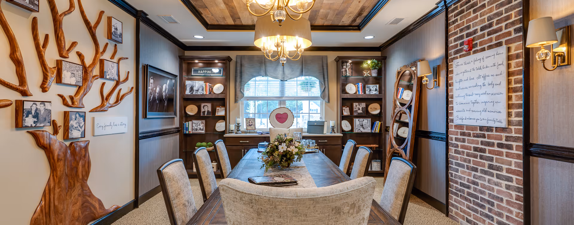 Well-decorated communal dining room with a long wooden table, upholstered chairs, chandelier, built-in shelves and decorative wall art including a wooden tree sculpture.