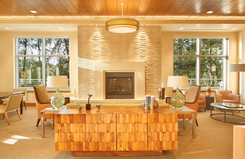 Bright, well-appointed lounge with a central wooden console, fireplace, armchairs, lamps, and large windows showing trees outside.
