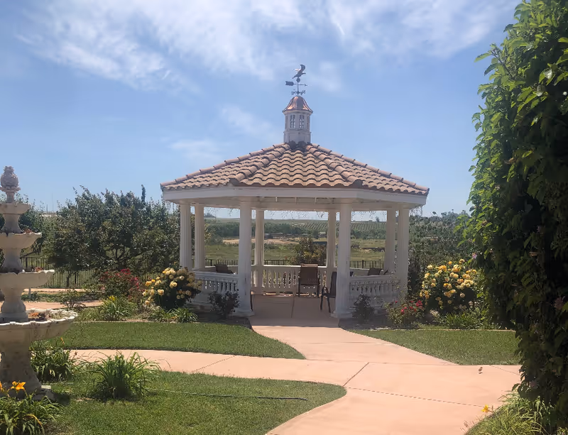 A white gazebo with a tiled roof and weather vane sits in a landscaped garden with pathways, flowers, and a fountain.