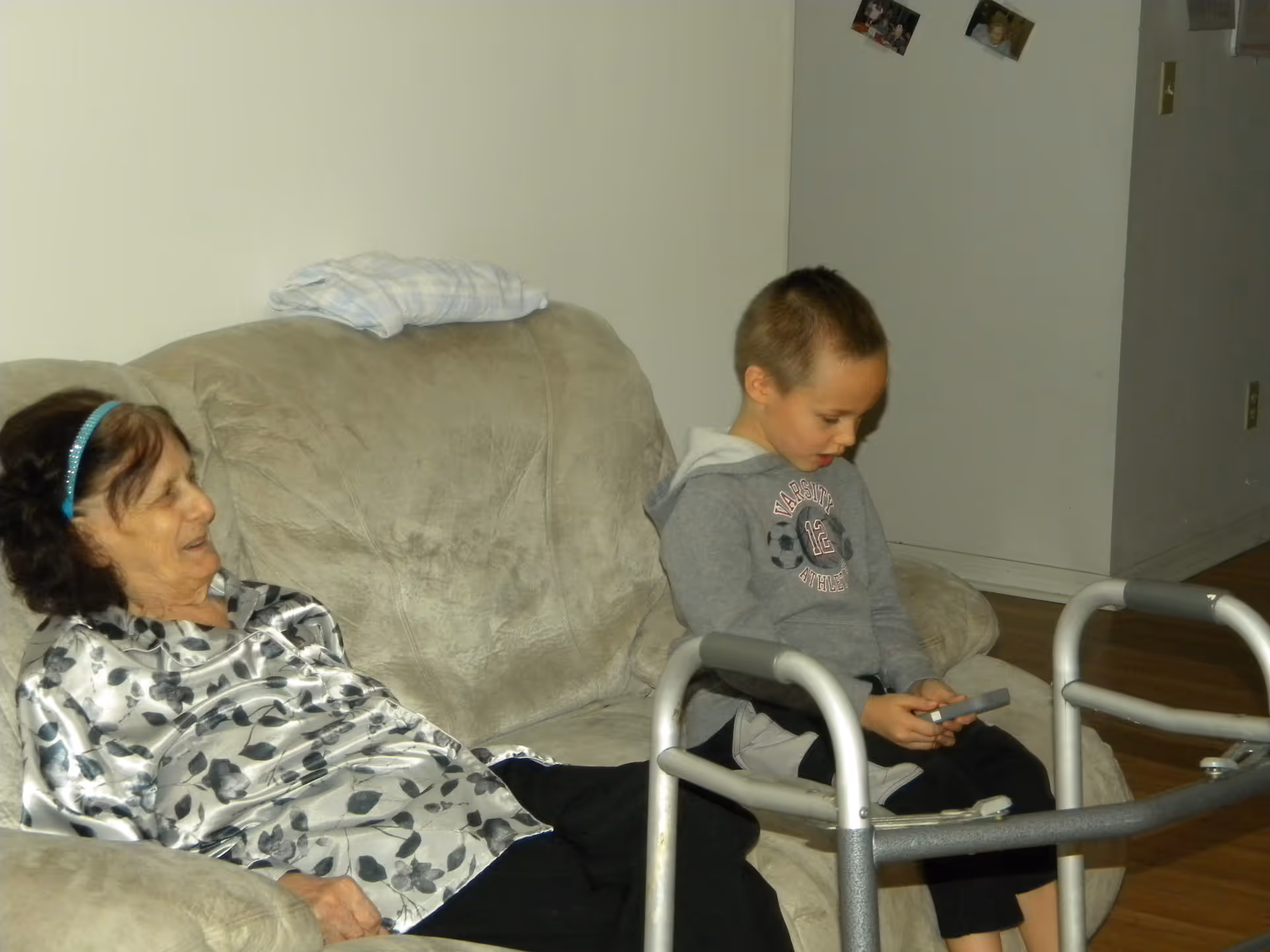 An elderly woman and a young boy sitting on a beige couch in a living room. The woman is wearing a floral patterned blouse and a blue headband, while the boy is wearing a gray hoodie and holding a remote control. A walker is positioned in front of the couch, and there are photos pinned on the wall in the background.