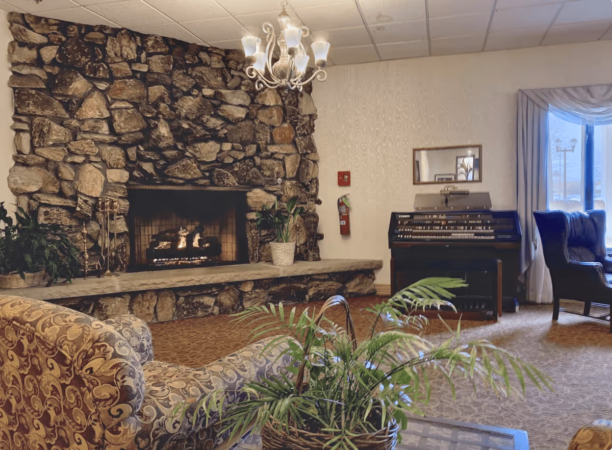 A cozy senior living facility common area with a large stone fireplace, a lit fire, and a stone hearth. There is a patterned upholstered sofa in the foreground, a green potted plant on a table, and another plant on the hearth. To the right, there is an organ with a mirror above it, a fire extinguisher mounted on the wall, a window with curtains, and a dark upholstered armchair.