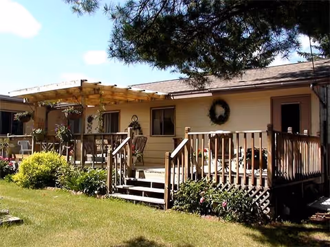 Single-story beige facility with wooden porch, railings, outdoor seating, and greenery.