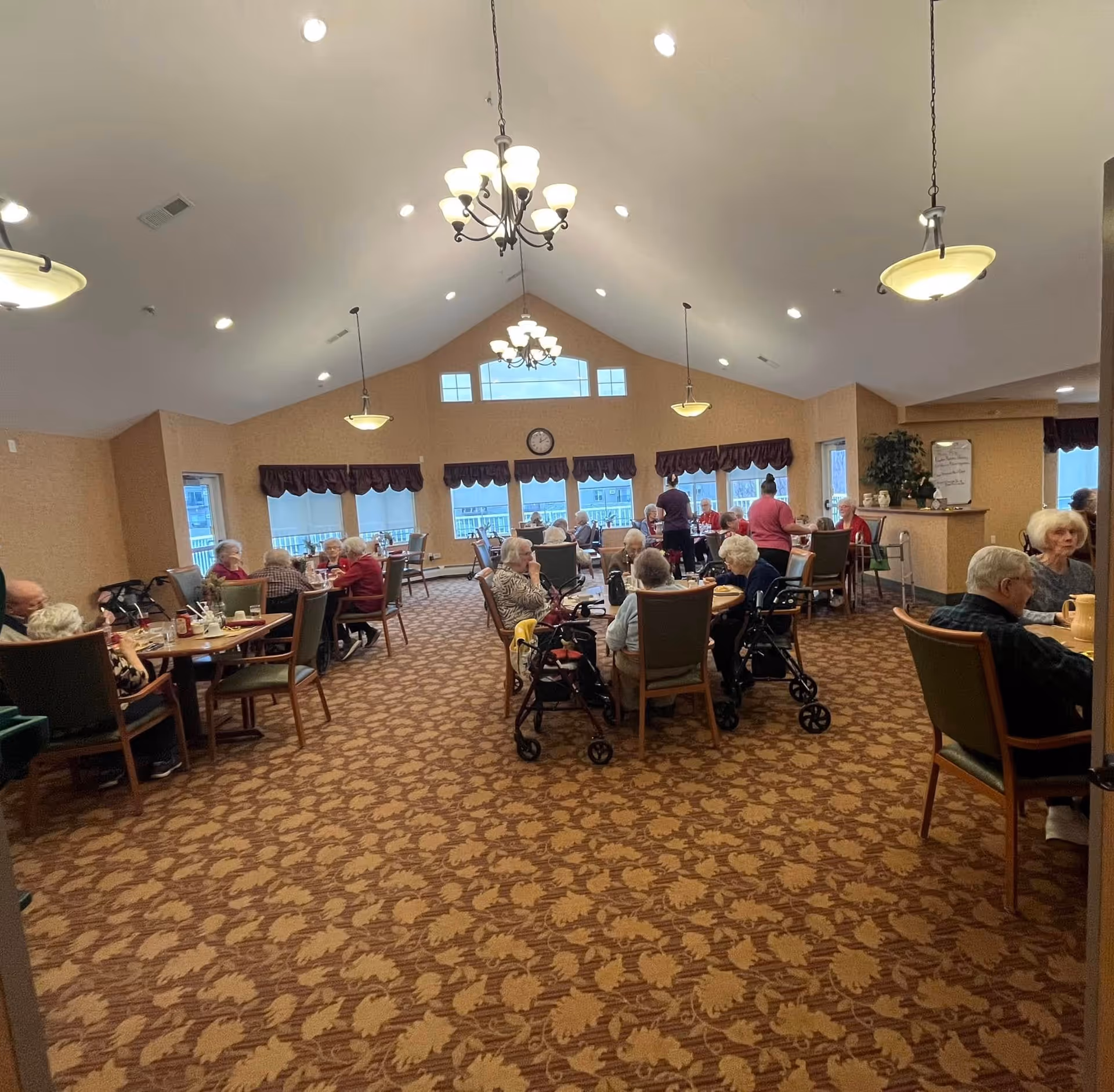 A large dining room in a senior living facility with many elderly residents seated at tables and staff assisting them.