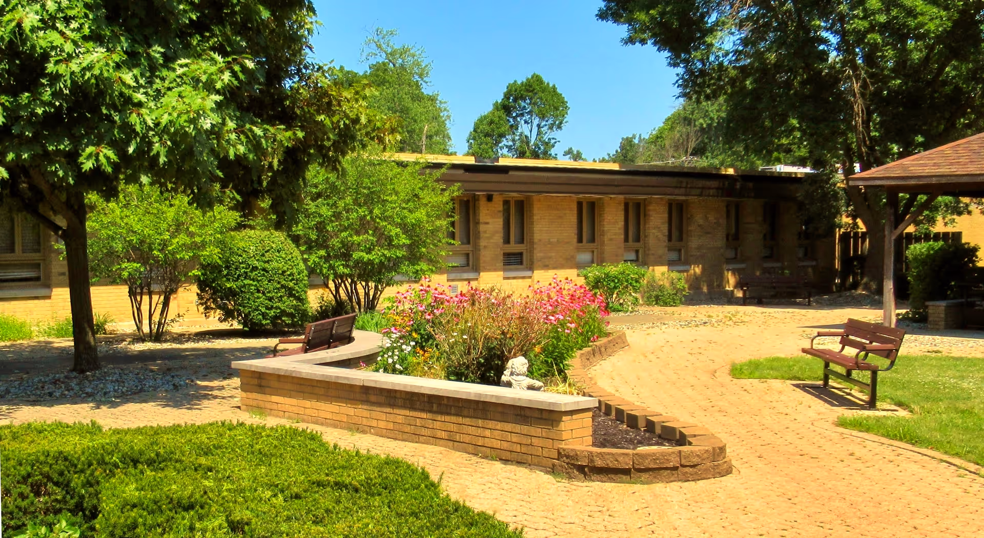 Outdoor courtyard area of a senior living facility with brick pathways, benches, flower beds, and trees surrounding a single-story brick building under a clear blue sky.