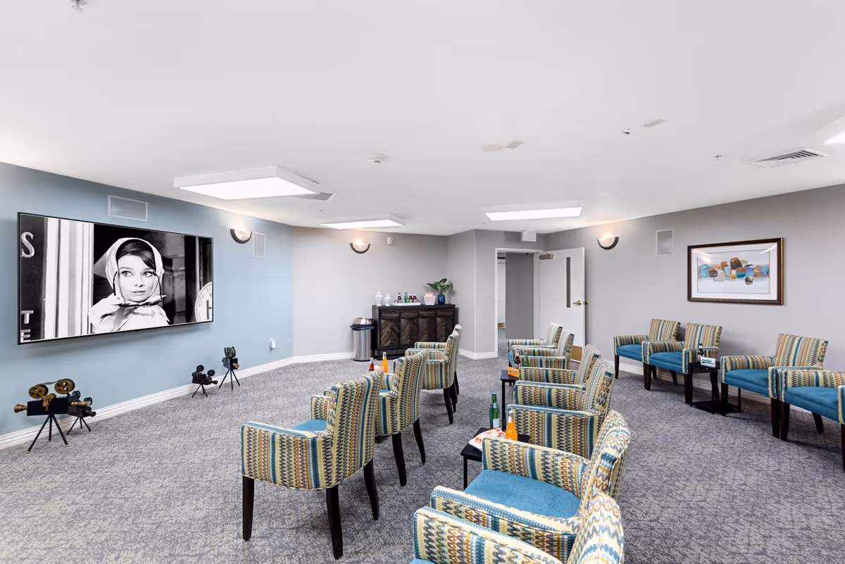 A senior living facility common room with patterned armchairs arranged in rows facing a large wall-mounted screen showing a black and white image of a woman. The room has gray carpet, light gray walls, modern ceiling lights, a sideboard with drinks and glasses, and framed artwork on the wall.