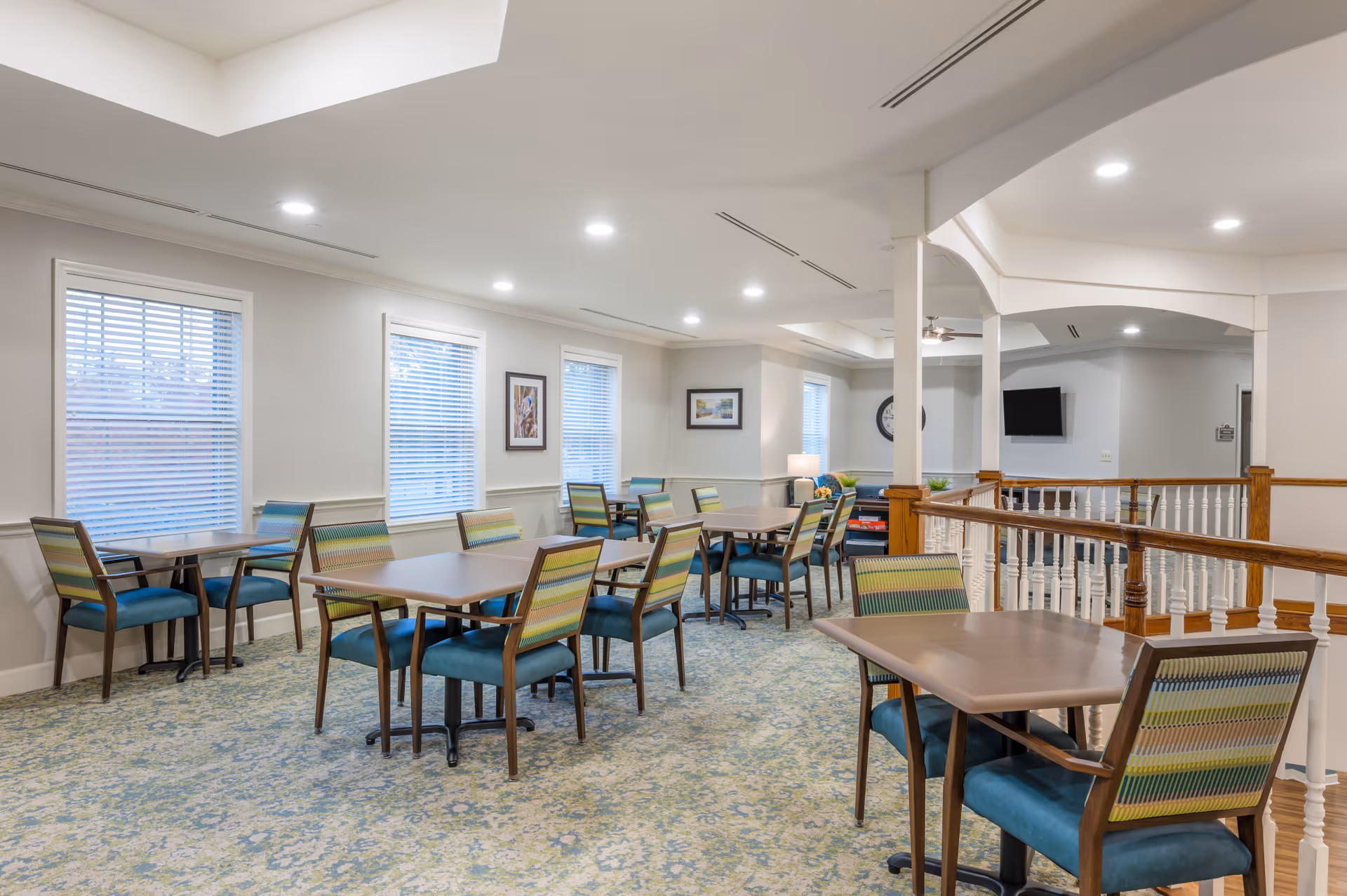 A bright and spacious dining area with multiple tables and chairs arranged neatly. The chairs have blue seats and patterned backs. The room features large windows with blinds, framed artwork on the walls, and a carpeted floor with a blue and green pattern. There is a wooden railing separating part of the room and a television mounted on the far wall.