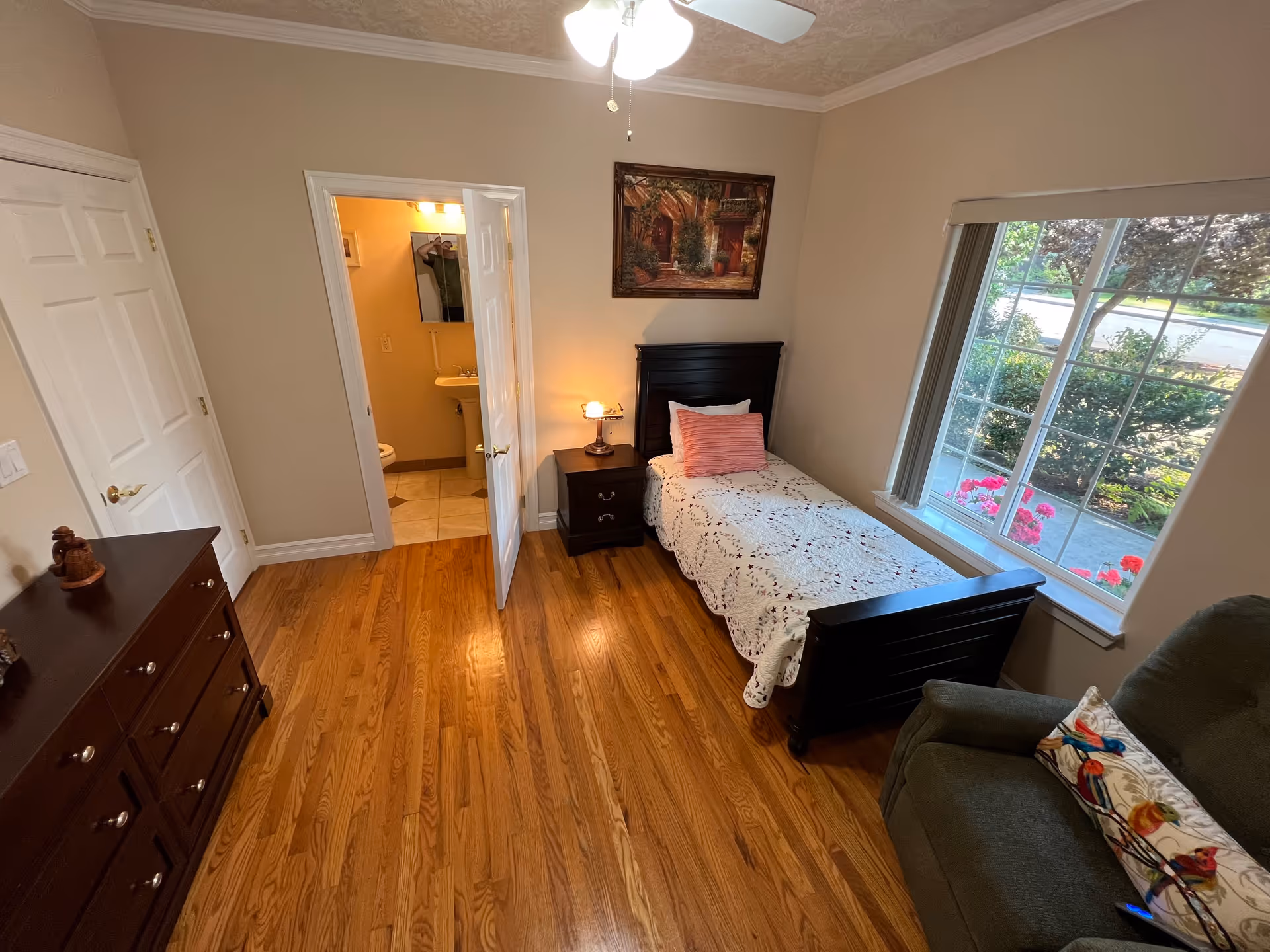 A small bedroom with a single bed covered with a white quilt and a pink pillow. Next to the bed is a dark wooden nightstand with a lamp. There is a large window with a view of greenery and flowers outside. A green armchair with a colorful bird-patterned pillow is positioned near the window. Across from the bed is a dark wooden dresser. The room has hardwood floors and beige walls. An open door reveals a small bathroom with a pedestal sink and toilet.