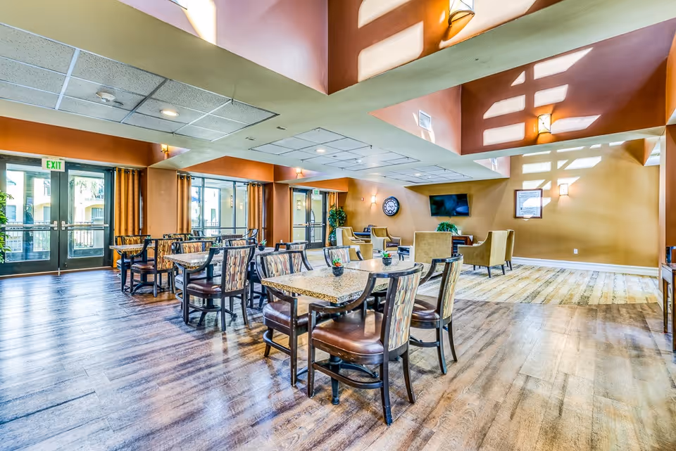 A spacious, well-lit common area with multiple tables and chairs arranged for dining or socializing. The room features large windows with curtains, a high ceiling with skylights, and a seating area with armchairs and a wall-mounted TV. The floor is wooden, and the walls are painted in warm tones.