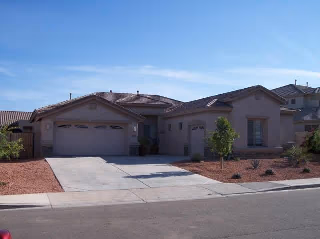 Single-story residential house with a three-car garage, beige exterior walls, and a tiled roof. The front yard has desert landscaping with red gravel, a few small plants, and a young tree. The sky is clear and blue.