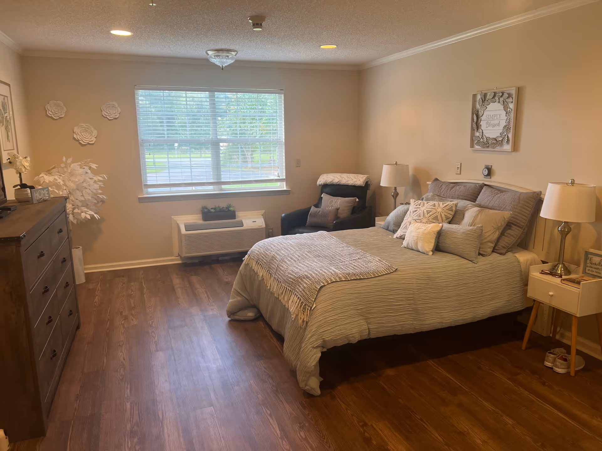 A cozy bedroom with a large bed covered in multiple pillows and a light gray bedspread. There is a dark armchair with a pillow and a blanket next to the bed. On either side of the bed are nightstands with lamps. A dresser with decorative items and a white artificial plant is on the left side of the room. The room has wooden flooring and a large window with blinds letting in natural light. The walls are decorated with framed pictures and wall art.