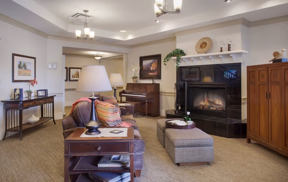 A cozy living room with a brown sofa adorned with colorful striped pillows and a pink throw blanket. There is a wooden side table with a lamp and a small tray on an ottoman in front of the sofa. The room features a black fireplace with a white mantel decorated with plants and ornaments. A wooden piano is placed against the wall, and framed artwork hangs above it. The room has beige carpet and neutral-colored walls, with additional lamps and furniture including a wooden cabinet and a console table with decorative items.