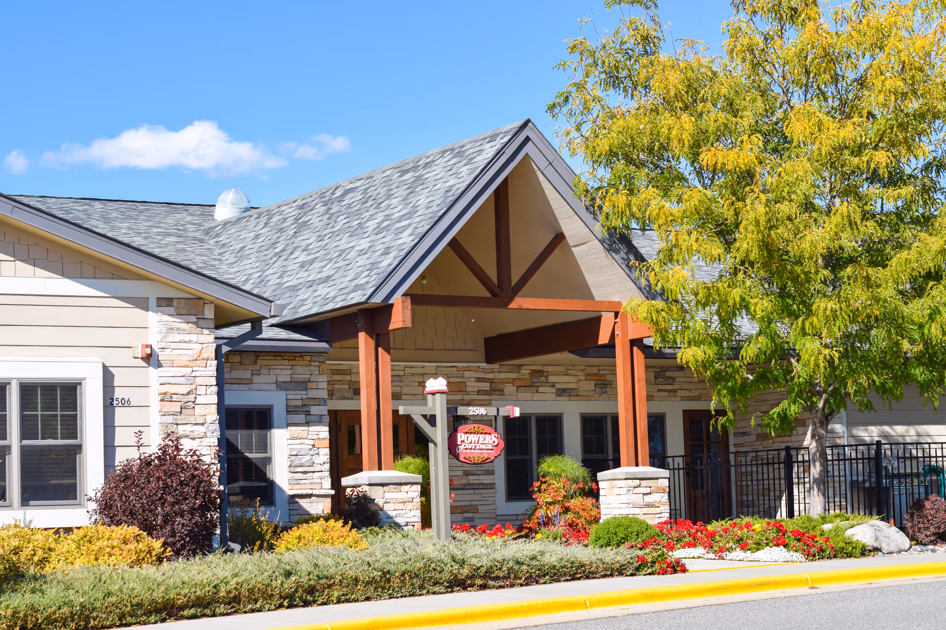 Front entrance of a single-story senior living building with a covered wooden portico, stone facade, and landscaped flowerbeds.