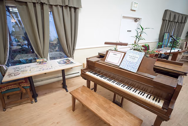 Interior room with a wooden piano and bench in the foreground, a table with puzzles and chairs near a window with curtains, and various plants and framed items on the piano.