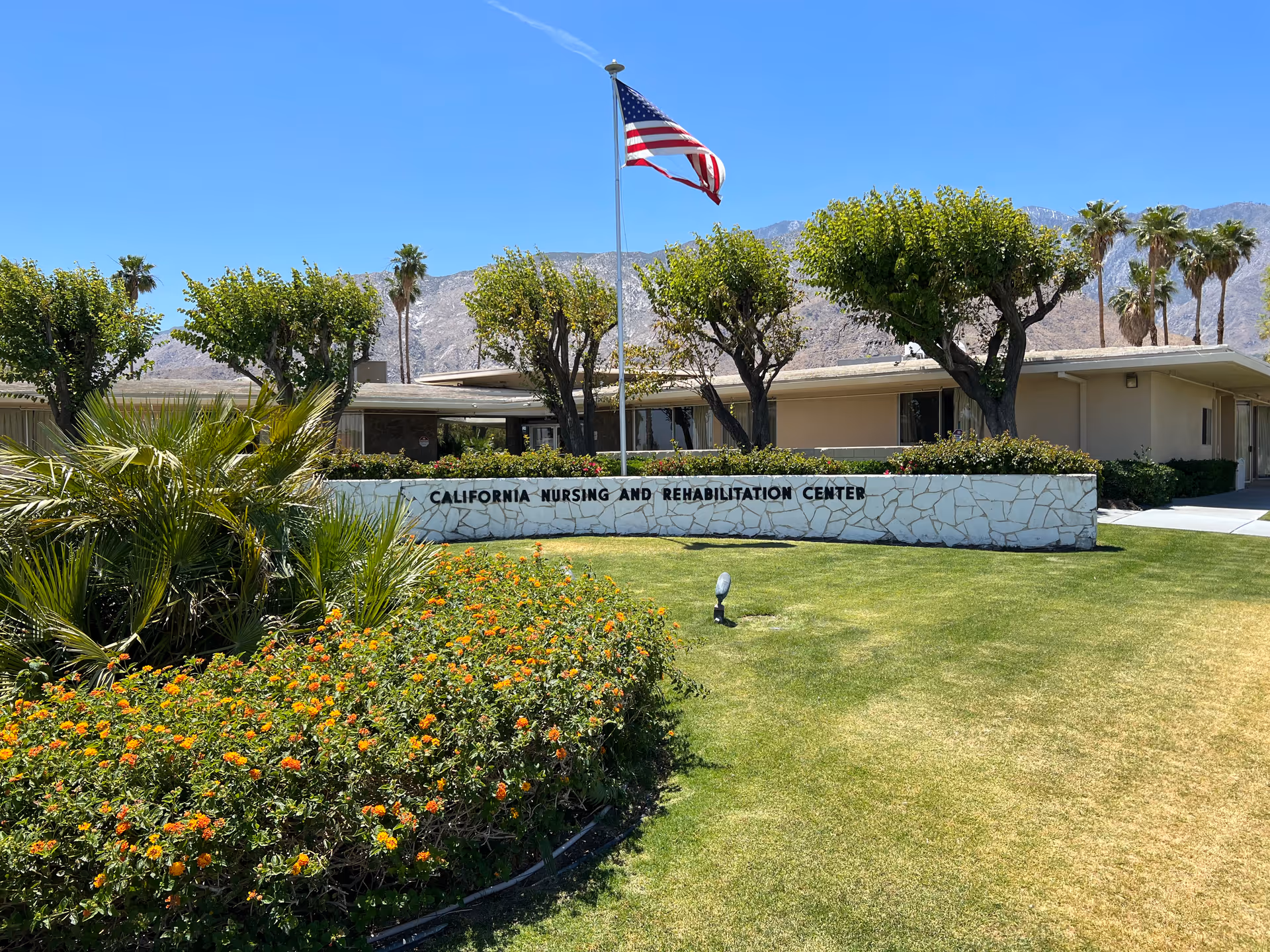 Exterior view of California Nursing and Rehabilitation Center building with a stone sign in front, an American flag on a flagpole, trimmed trees, flowering bushes, and mountains in the background under a clear blue sky.