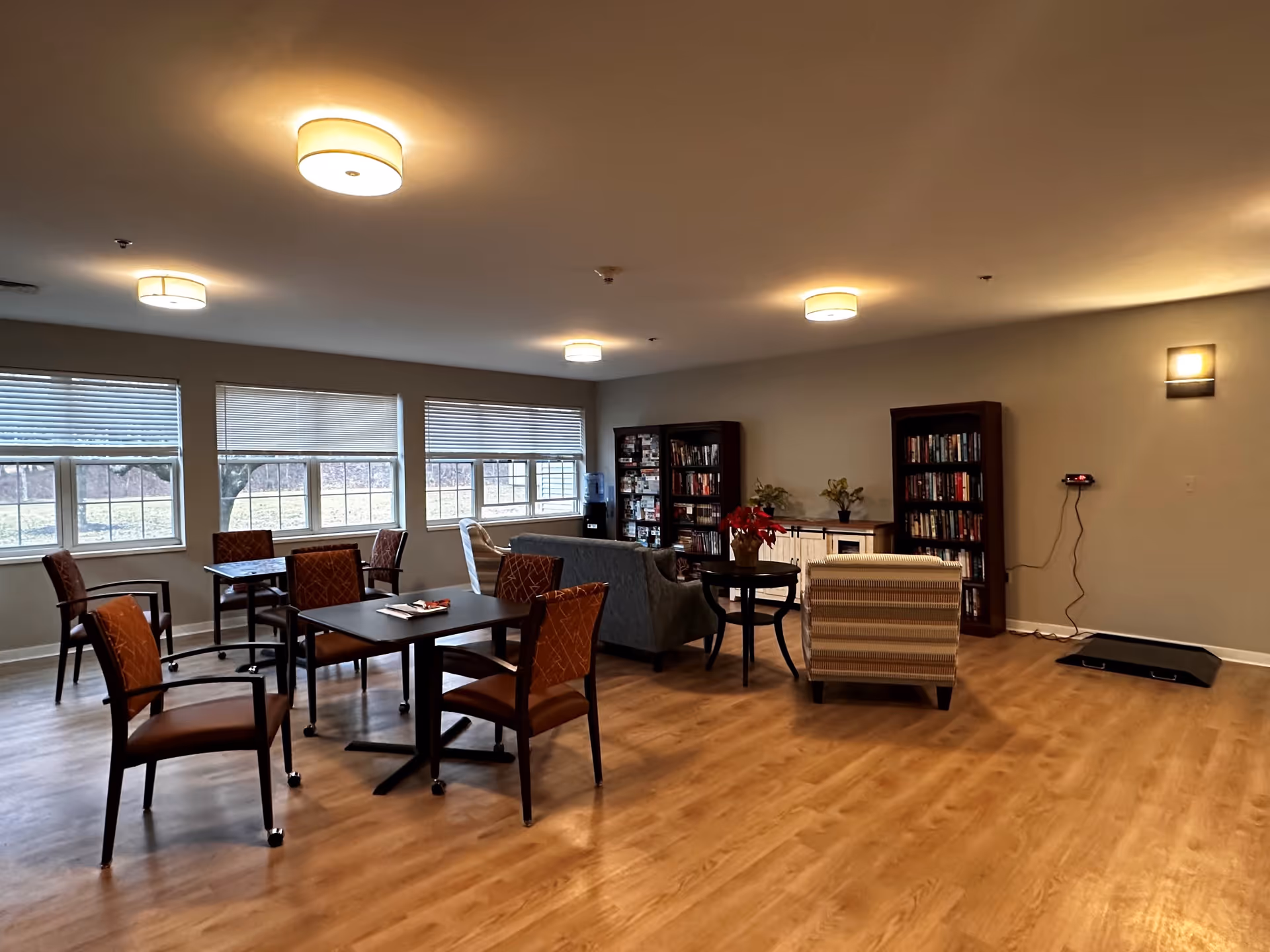 A spacious common area in a senior living facility with several tables and chairs arranged near large windows. There are two bookshelves filled with books, a water dispenser, a small cabinet with plants and flowers on top, and comfortable armchairs. The room has wooden flooring and ceiling lights providing warm illumination.