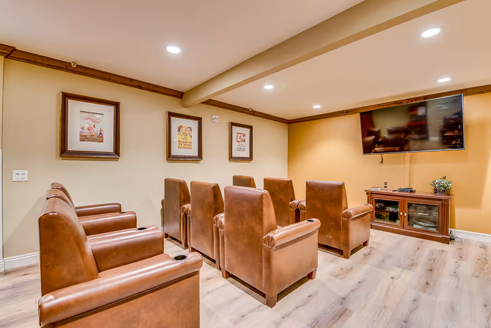 A small media room with two rows of brown leather recliner chairs facing a wall-mounted TV and wooden media cabinet.