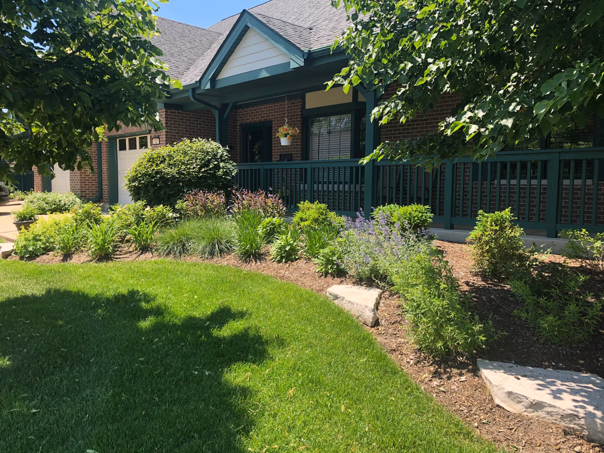 Front exterior view of a brick building with green trim, a covered porch with railing, hanging flower pot, and well-maintained garden with various shrubs and plants in front of a green lawn under a sunny sky.