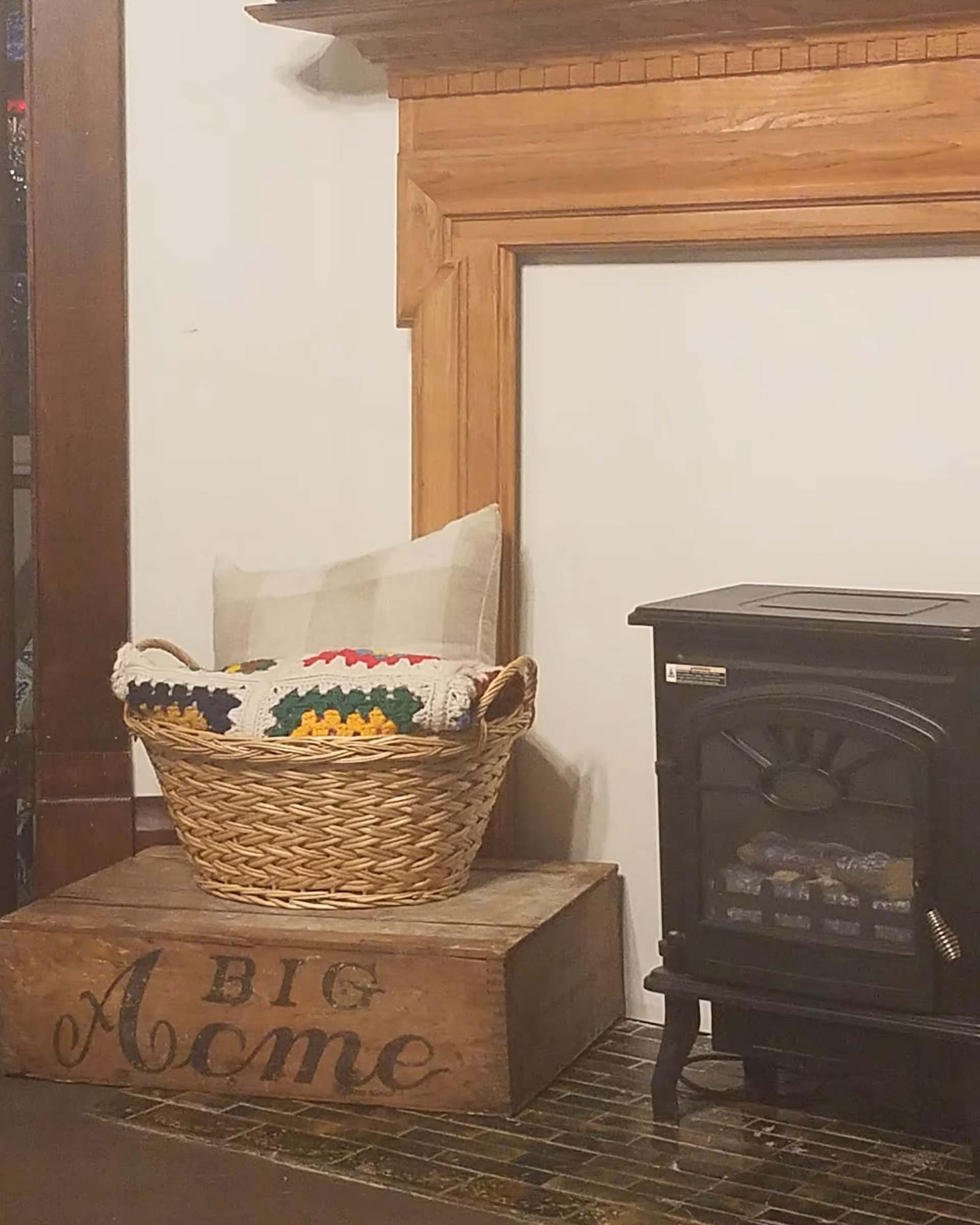 A cozy corner featuring a small black electric stove heater next to a wooden mantel. On the left side, there is a wooden box with the words 'A BIG Acme' painted on it, holding a wicker basket filled with a plaid pillow and a colorful crocheted blanket.