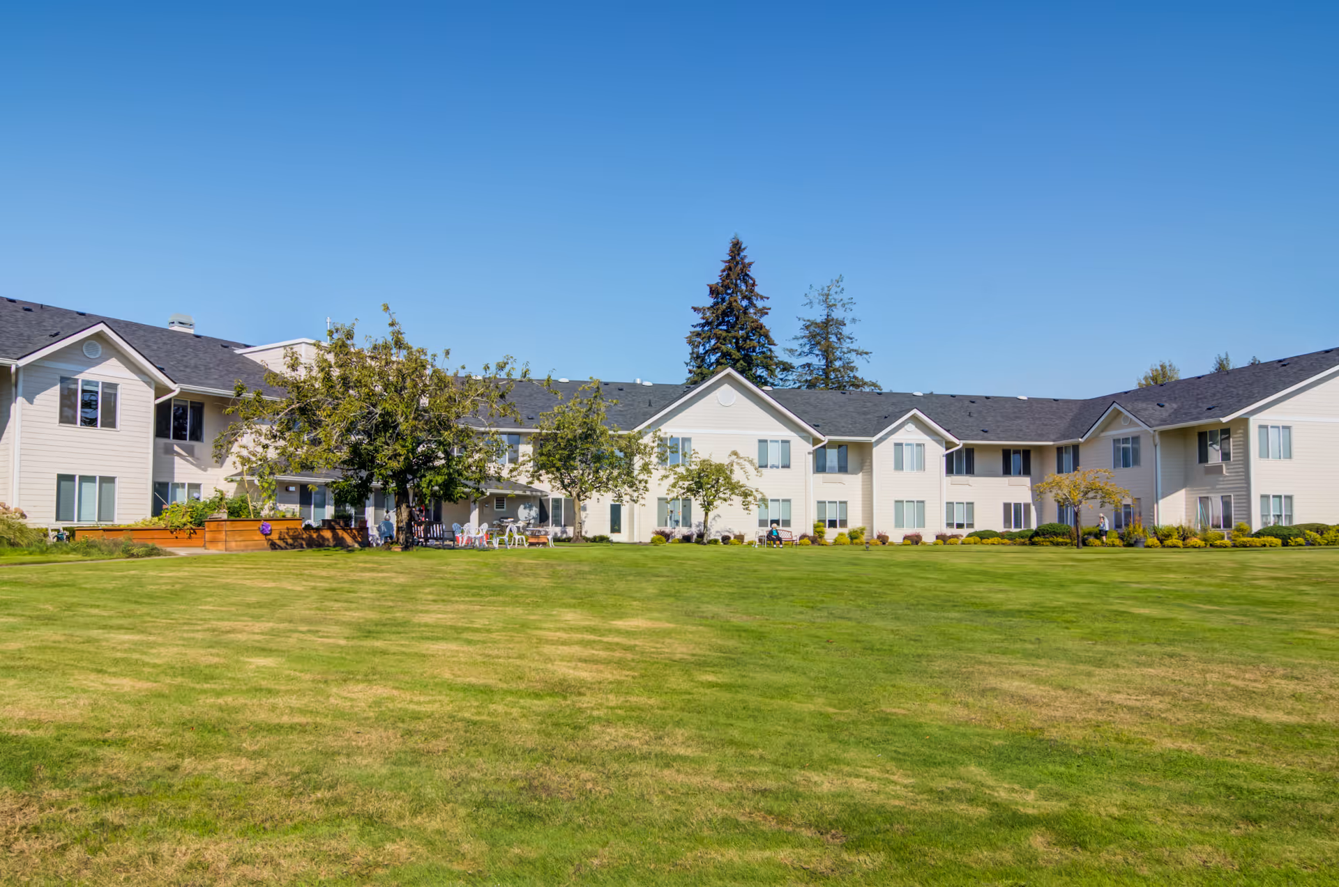 Wide exterior view of a two-story assisted living facility building with beige siding and a dark roof, surrounded by a large green lawn and several trees under a clear blue sky.